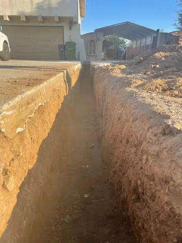 Deep open trench beside a house driveway with excavated soil nearby