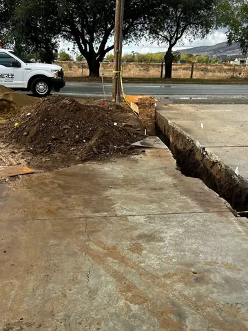 Open trench beside a driveway and front yard with excavated soil and a street beyond