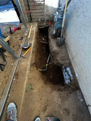 Narrow trench along a house wall with conduit and tools; shoes appear at the upper edge