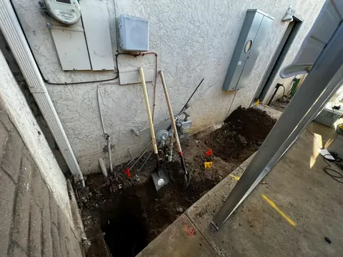 Open trench beside a house wall with nearby electrical enclosures and conduit